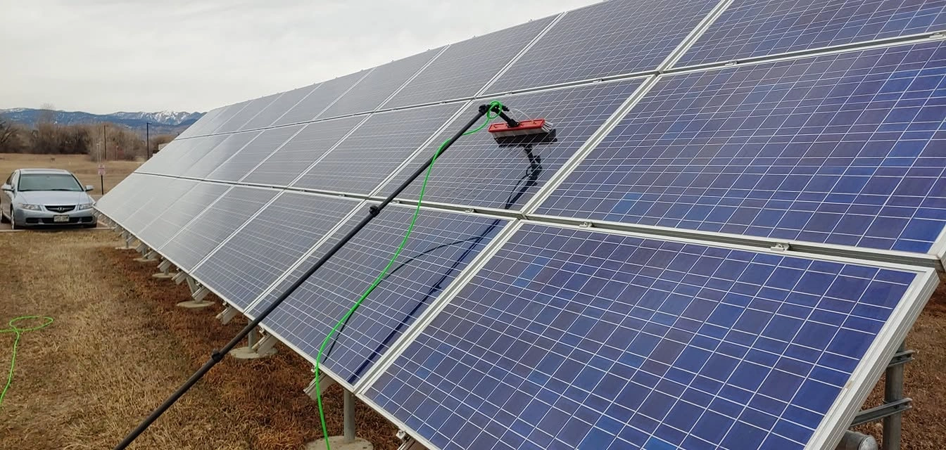 A water-fed extension pole and soft brush assembly cleaning a ground-mounted solar panel array